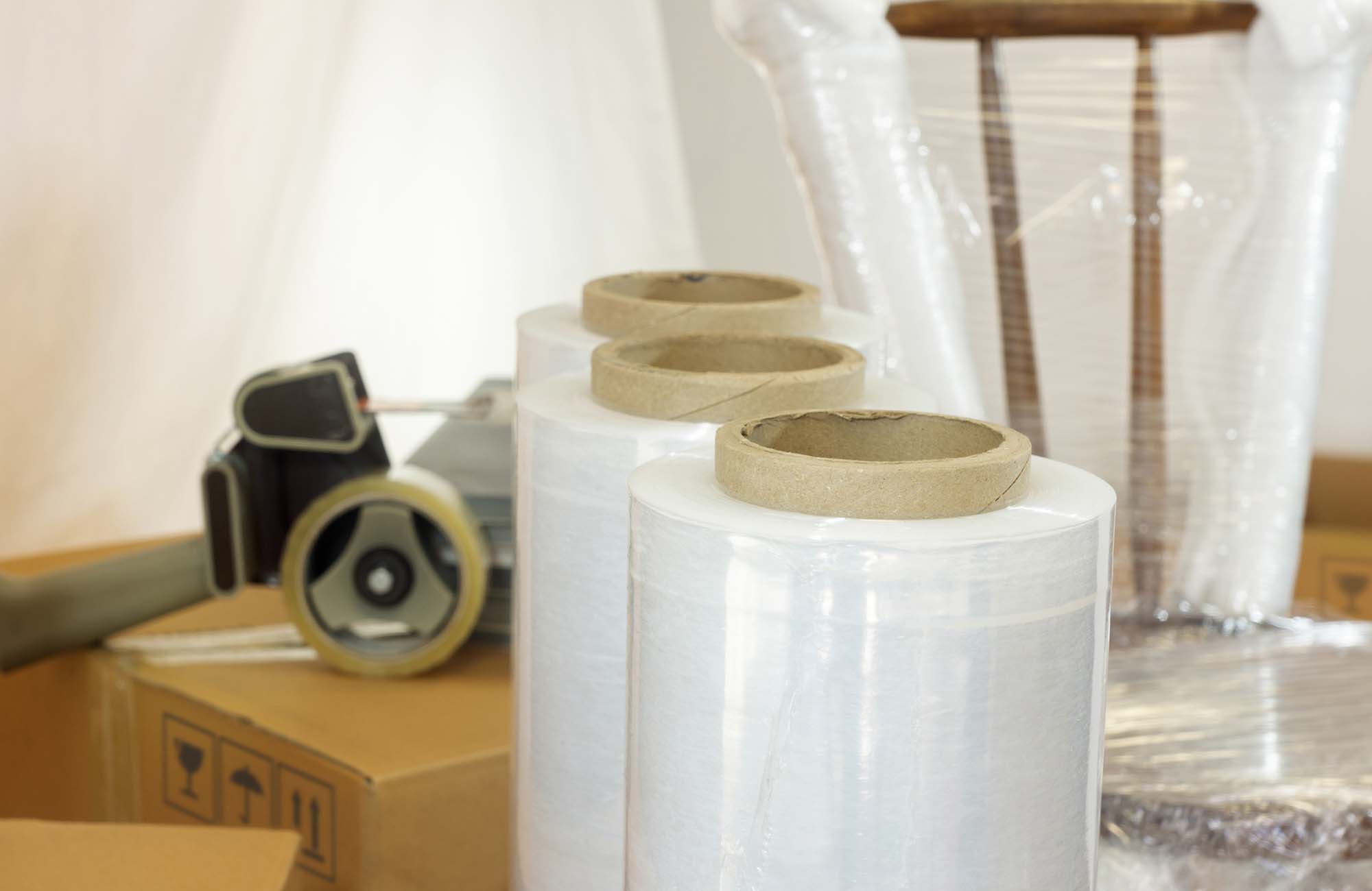 Approach to plastic rolls for packing in the foreground with a plastic wrapped chair, cardboard boxes and adhesive